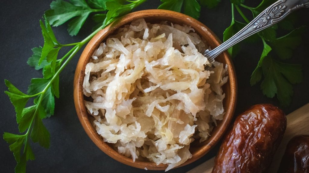 From above, a small wooden bowl is filled with tangy sauerkraut, its textured appearance offering a hint of its sharp flavor. A metal teaspoon rests adjacent, ready for serving. This setting highlights sauerkraut as a quintessential garnish, perfect as a bratwurst or hotdog topping, adding a delightful crunch and zest to each bite.