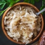 From above, a small wooden bowl is filled with tangy sauerkraut, its textured appearance offering a hint of its sharp flavor. A metal teaspoon rests adjacent, ready for serving. This setting highlights sauerkraut as a quintessential garnish, perfect as a bratwurst or hotdog topping, adding a delightful crunch and zest to each bite.